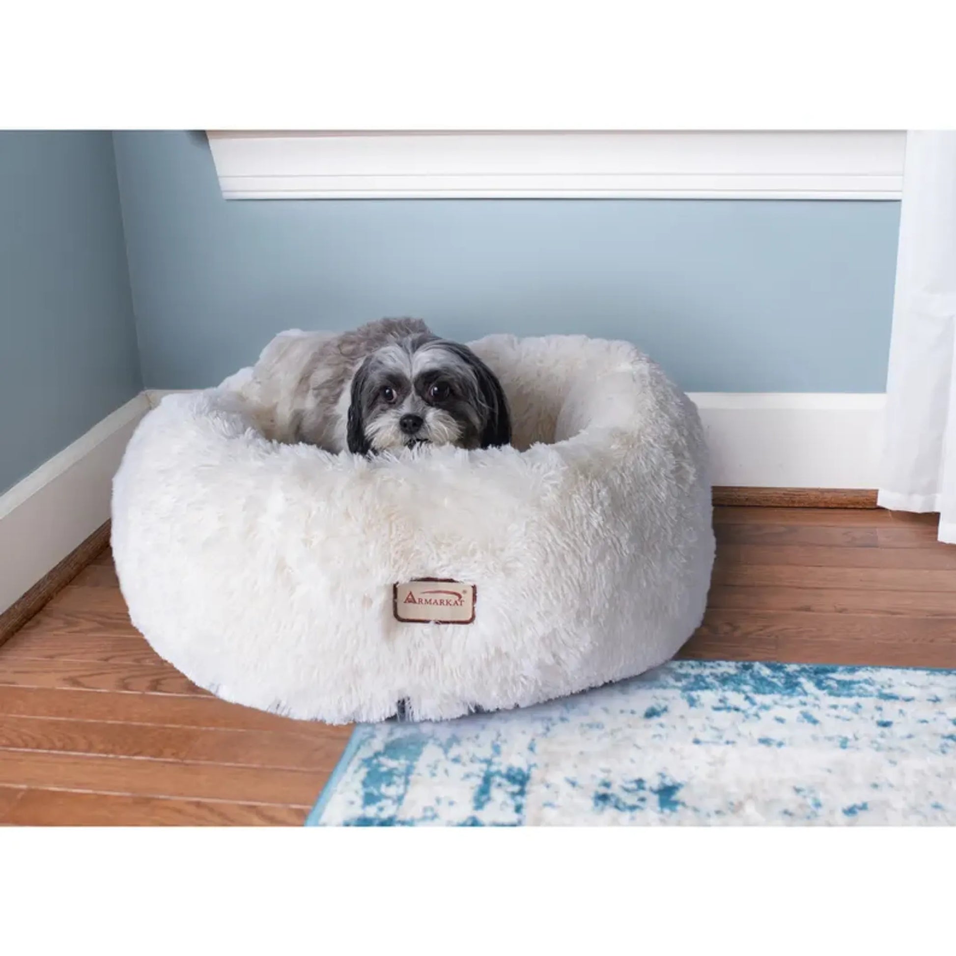 Dog lying in a fluffy white pet bed with a visible brand logo on a wooden floor.